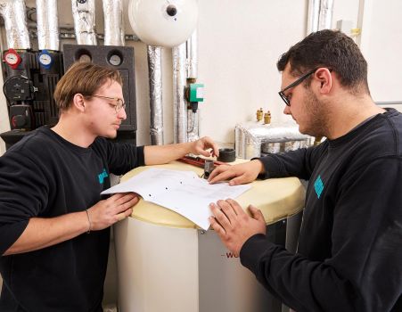 Zwei Mitarbeitende von WEME Heizung & Sanitär prüfen gemeinsam Pläne an einer Heizungsanlage im Technikraum – Zusammenarbeit im SHK-Team