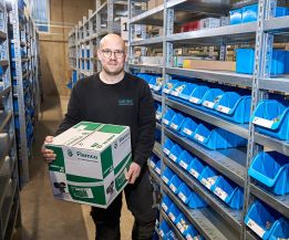 Mitarbeiter von WEME Heizung & Sanitär steht im Lager zwischen Regalen mit blauen Sichtboxen und hält einen Karton mit Heizungs- und Sanitärkomponenten
