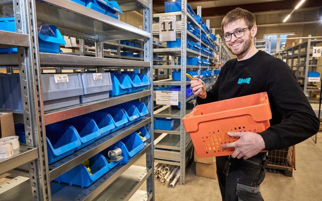 Mitarbeiter von WEME Heizung & Sanitär im Lager, steht vor Regalen mit sortierten Kleinteilen, hält einen orangefarbenen Materialkorb und prüft ein Bauteil
