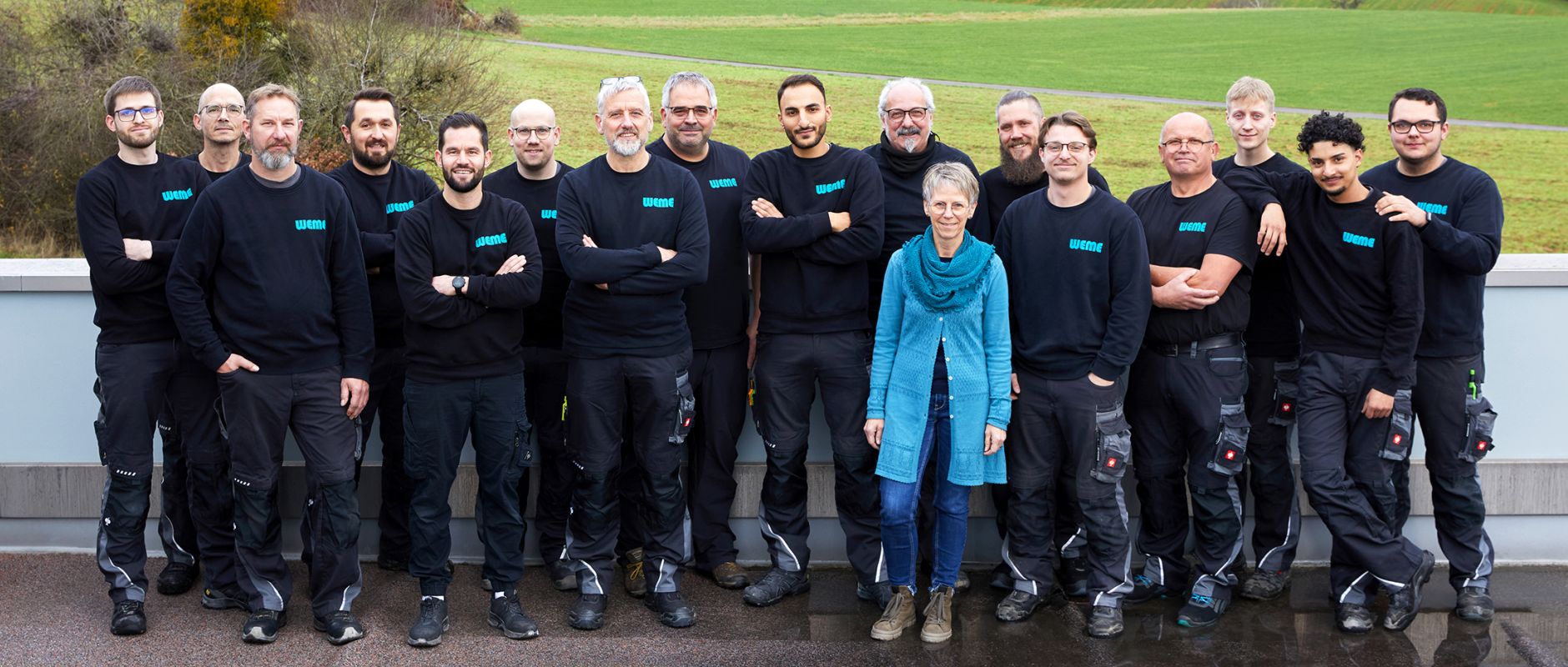 Gruppenfoto des gesamten WEME Heizung & Sanitär Teams in Arbeitskleidung, Mitarbeitende stehen gemeinsam im Freien vor einer grünen Landschaft