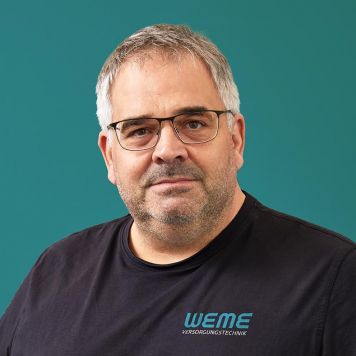 Geschäftsführer Alexander Puderbach von WEME Versorgungstechnik steht vor petrolfarbenem Hintergrund, trägt schwarze Arbeitskleidung mit WEME-Logo