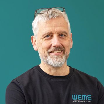Geschäftsführer Ralf Meyer von WEME Versorgungstechnik steht vor petrolfarbenem Hintergrund, trägt schwarze Arbeitskleidung mit WEME-Logo